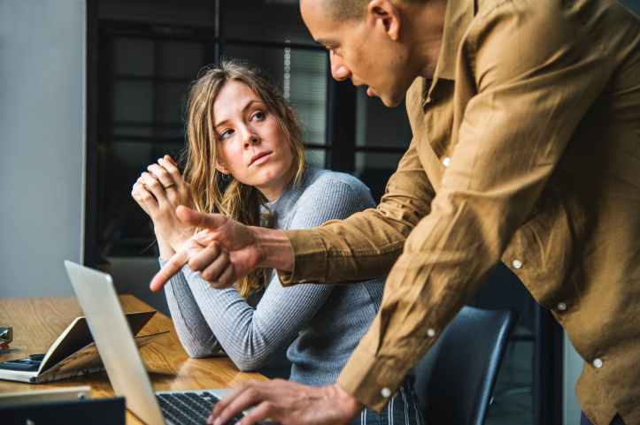man in brown long sleeved button up shirt standing while using gray laptop computer on brown wooden table beside woman in gray long sleeved shirt sitting